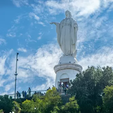 A huge statue of the Virgin Mary at the top of a hill in a city park, with people standing at an overlook in front.