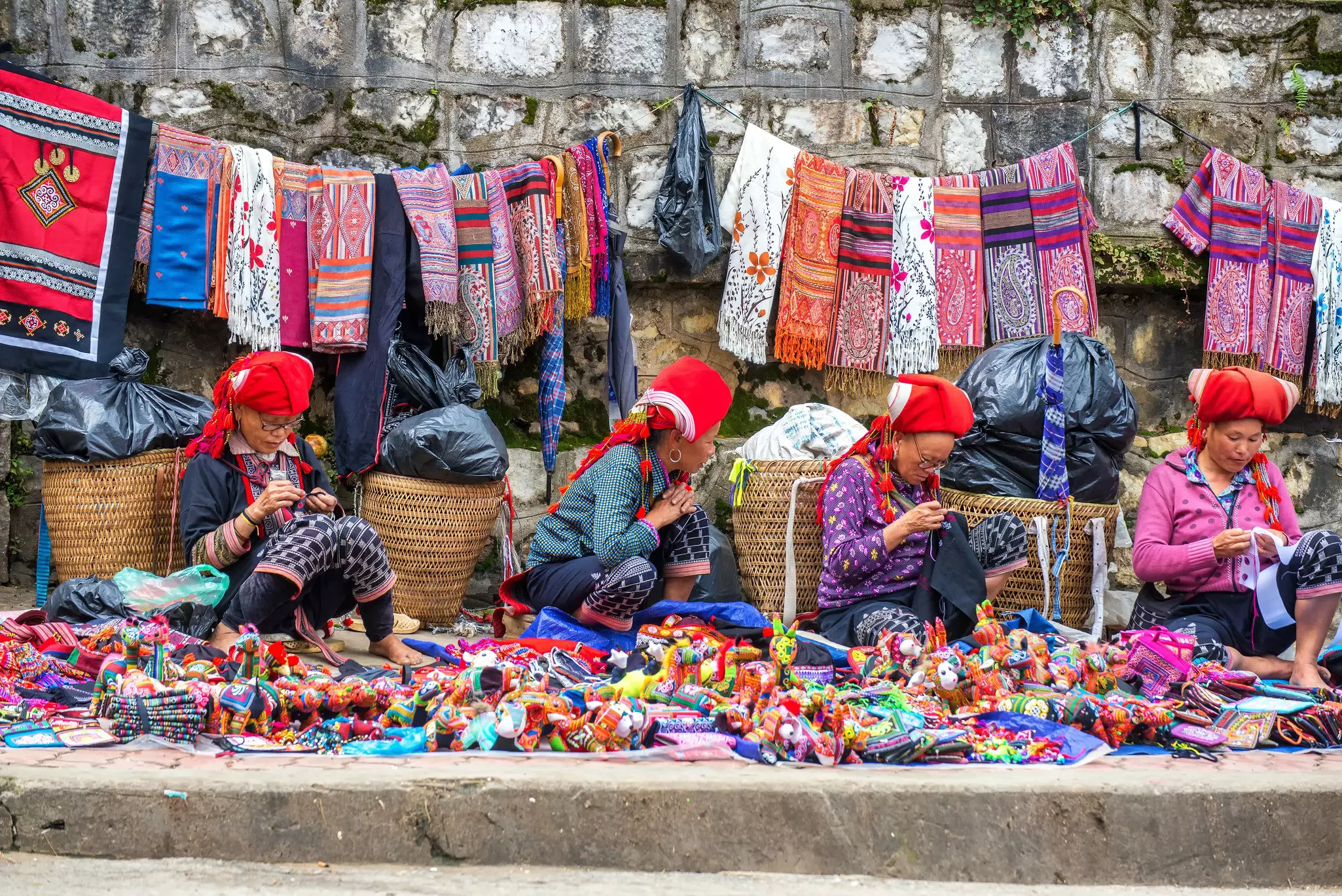 Four Hmong women sit in front of baskets as they work on handcrafts. Traditional woven fabrics hang on the rock wall behind them, and more goods are spread in front of them on the main street in Sapa, Vietnam