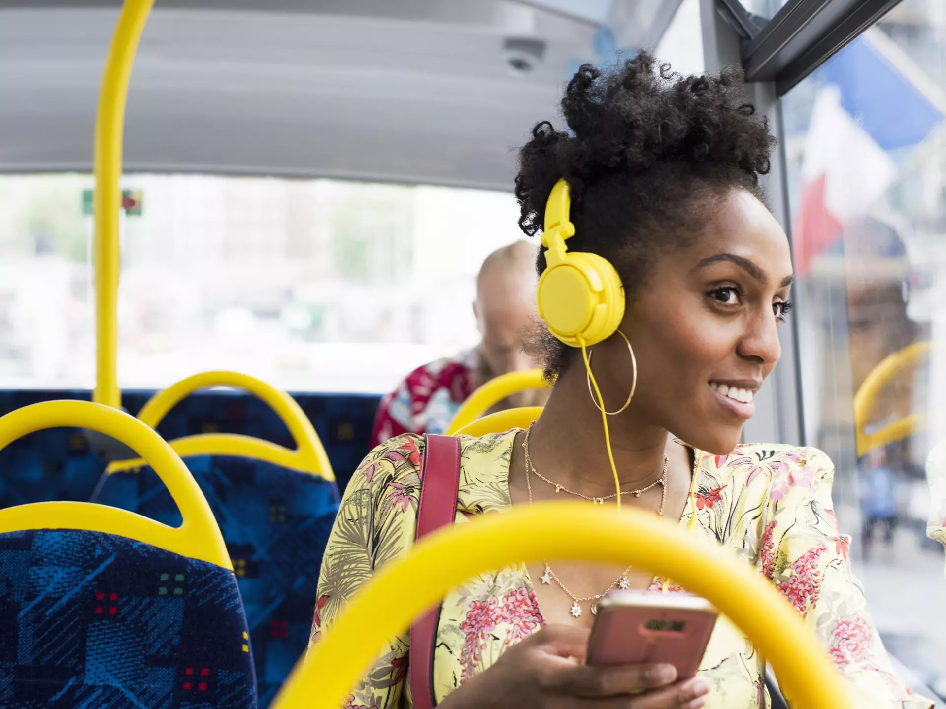 A black woman wearing headphones is looking out of the window of a bus and smiling