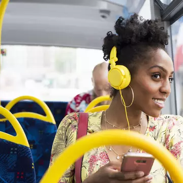 A black woman wearing headphones is looking out of the window of a bus and smiling