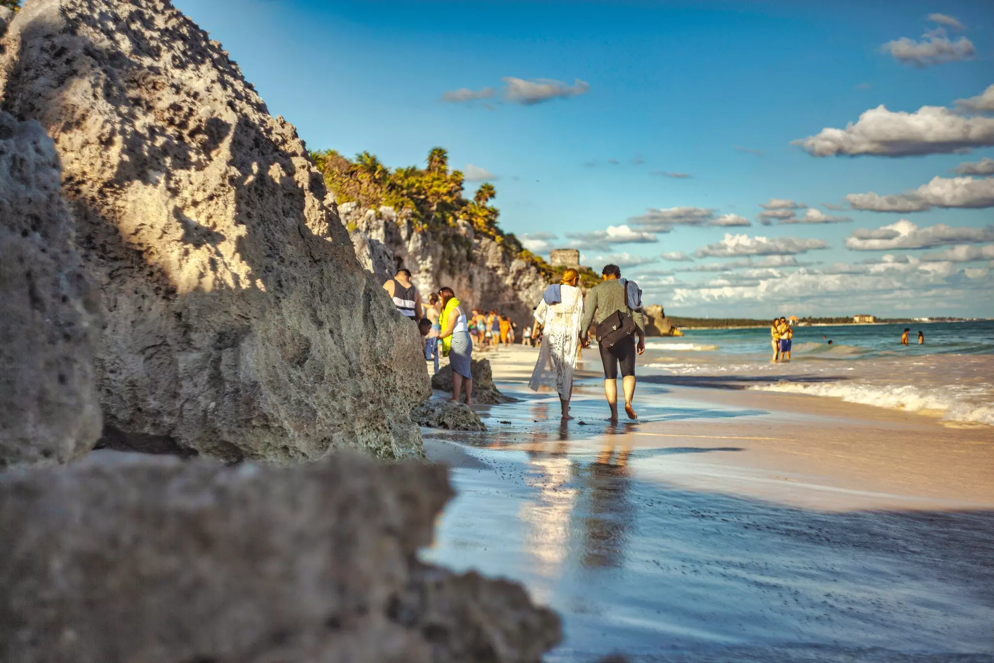 A couple walk arm in arm along a sandy beach with rocks to one side as people splash in the ocean