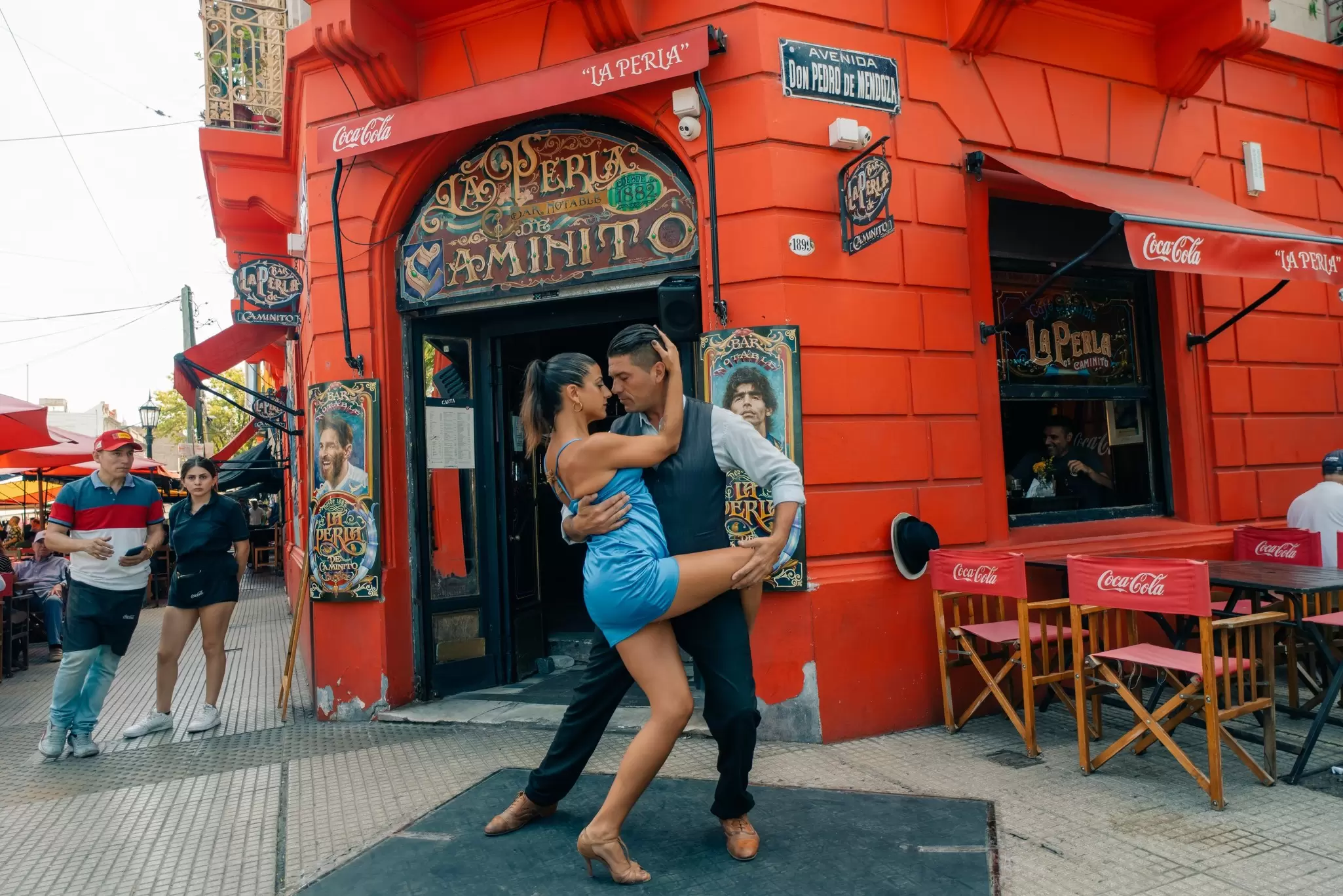 Tango dancers in a close pose on a sidewalk in front of a red building.