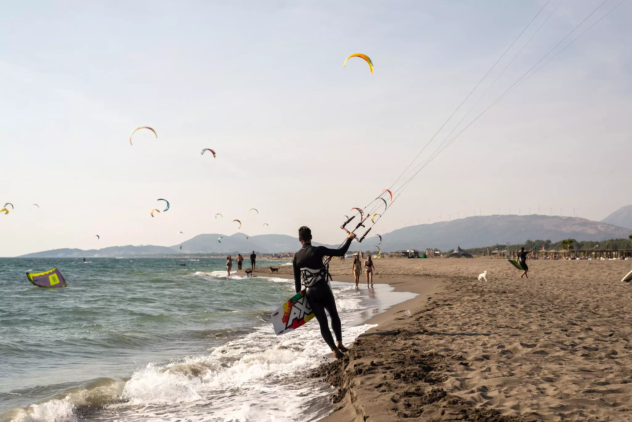 A kitesurfer holds a board in one hand and a kite handle in the other at the edge of a sandy beach in Montenegro.