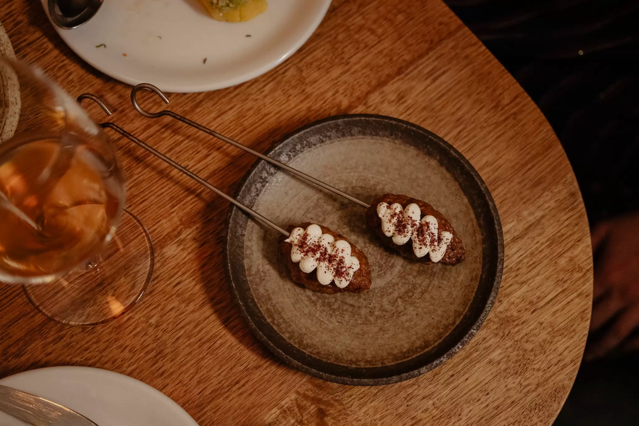 Food on skewers on a plate placed on a wooden table. A wine glass is nearby.