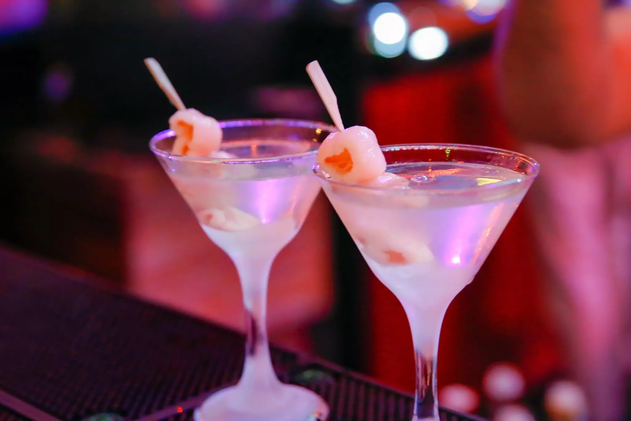 A trio of women with their backs facing the camera stand in the middle of a field, facing the sun. One woman is raising a wine glass, another is raising a wine bottle and another is holding a bouquet of flowers; bachelorette party