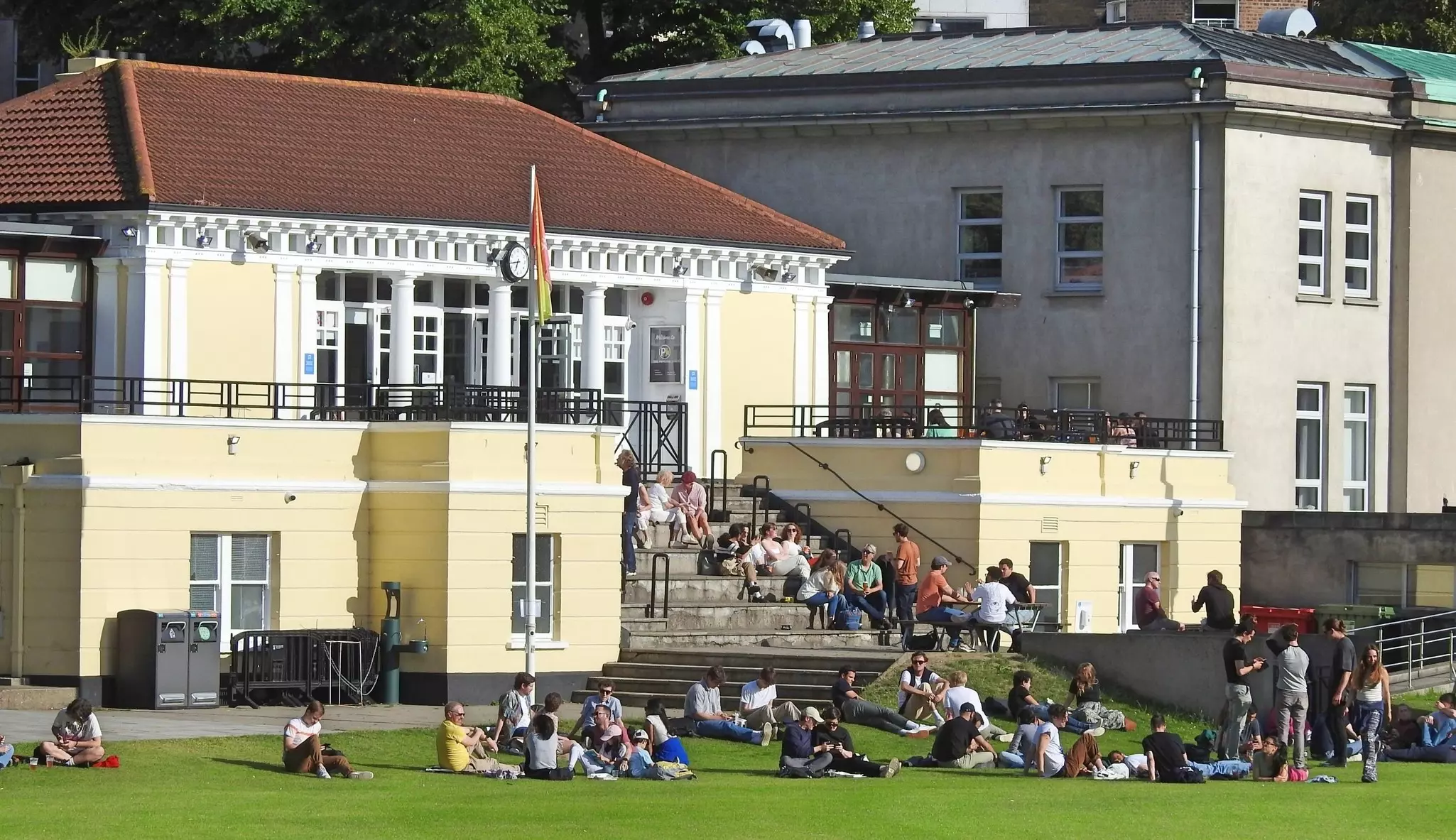 People lie on the grass of a sports field in front of a pavilion with a cafe.