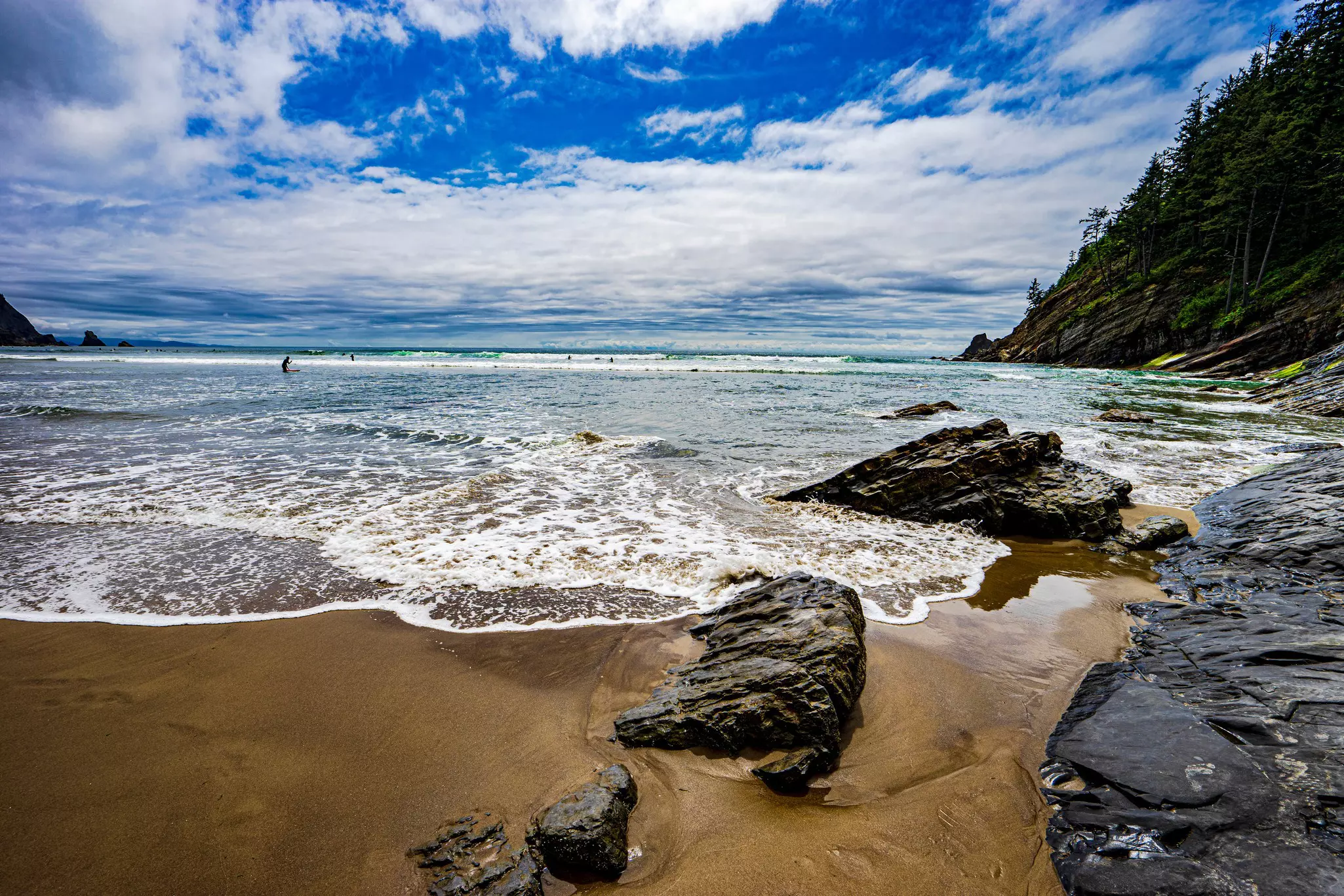Waves lapping at rocks on a sandy beach. Surfers await waves out at sea.