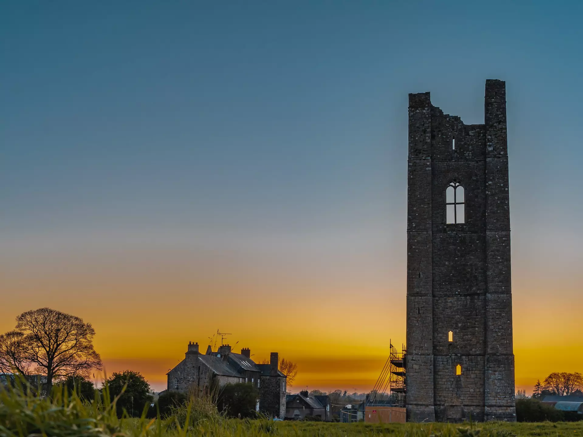A golden yellow sunset with a stone tower, stone house, and leafless tree in the foreground.