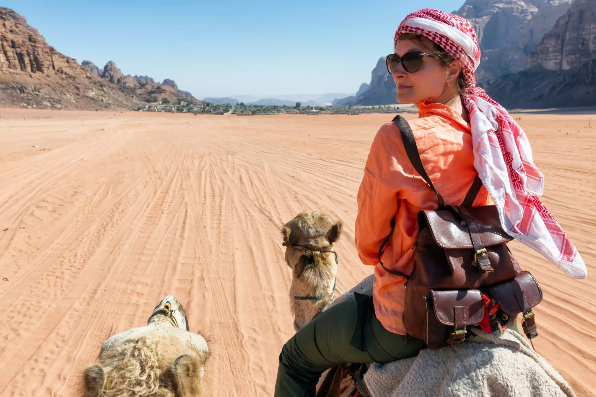 A person riding a camel wearing a head wrap looks over their left shoulder; the landscape ahead is reddish sand with rock formations on the perimeter.