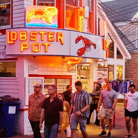 People walking past a restaurant with a neon sign that says "Lobster Pot" and a red lobster logo above the doorway.