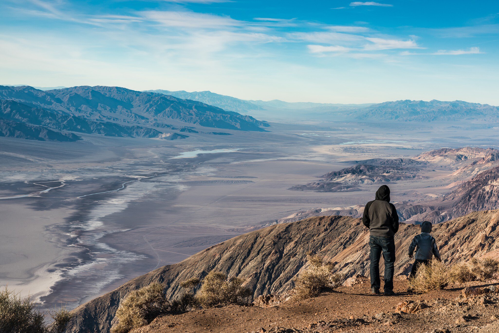 Two hooded figures seen from behind on a rocky ridge edge above white salt flats and a broad valley ringed by mountains