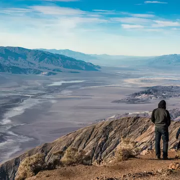 Two hooded figures seen from behind on a rocky ridge edge above white salt flats and a broad valley ringed by mountains