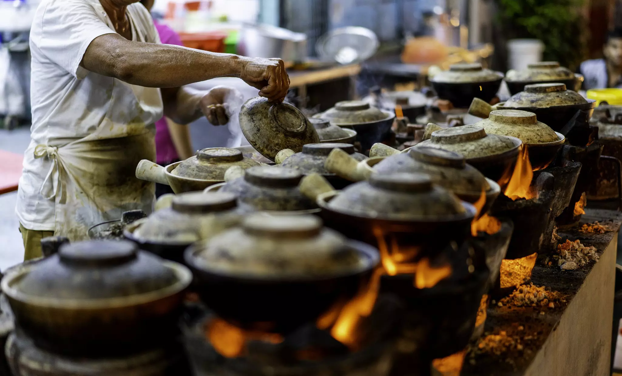 A vendor in Malaysia stands in front of many clay pots sitting over flames.