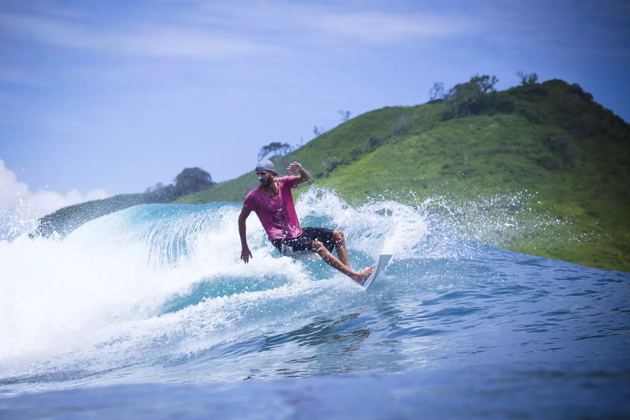 A surfer wearing a pink shirt and a blue cap rides a surfboard with a wave crashing behind him.