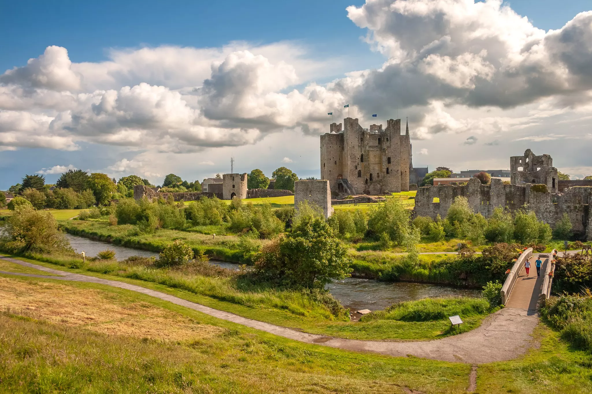 A gray castle surrounded by high bushes and trees. Two people walk a bridge over water away from it.