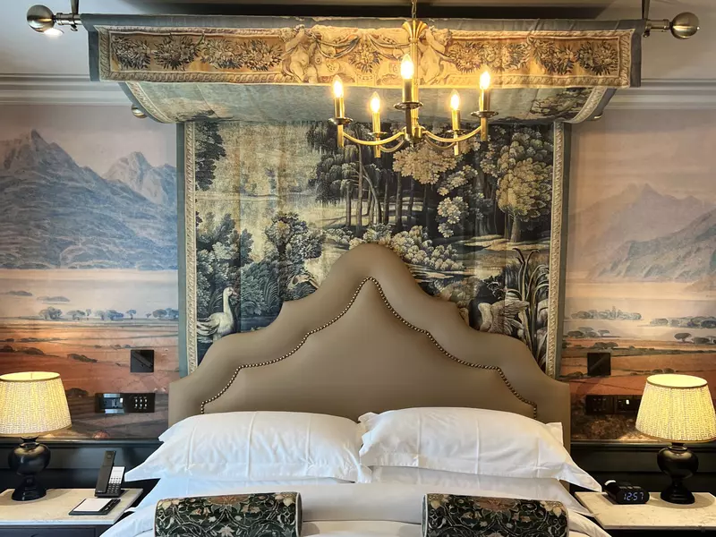 A bed with a white covering and round patterned pillows; the triangular headboard is beige, and the wall behind it has layered pieces of artwork.