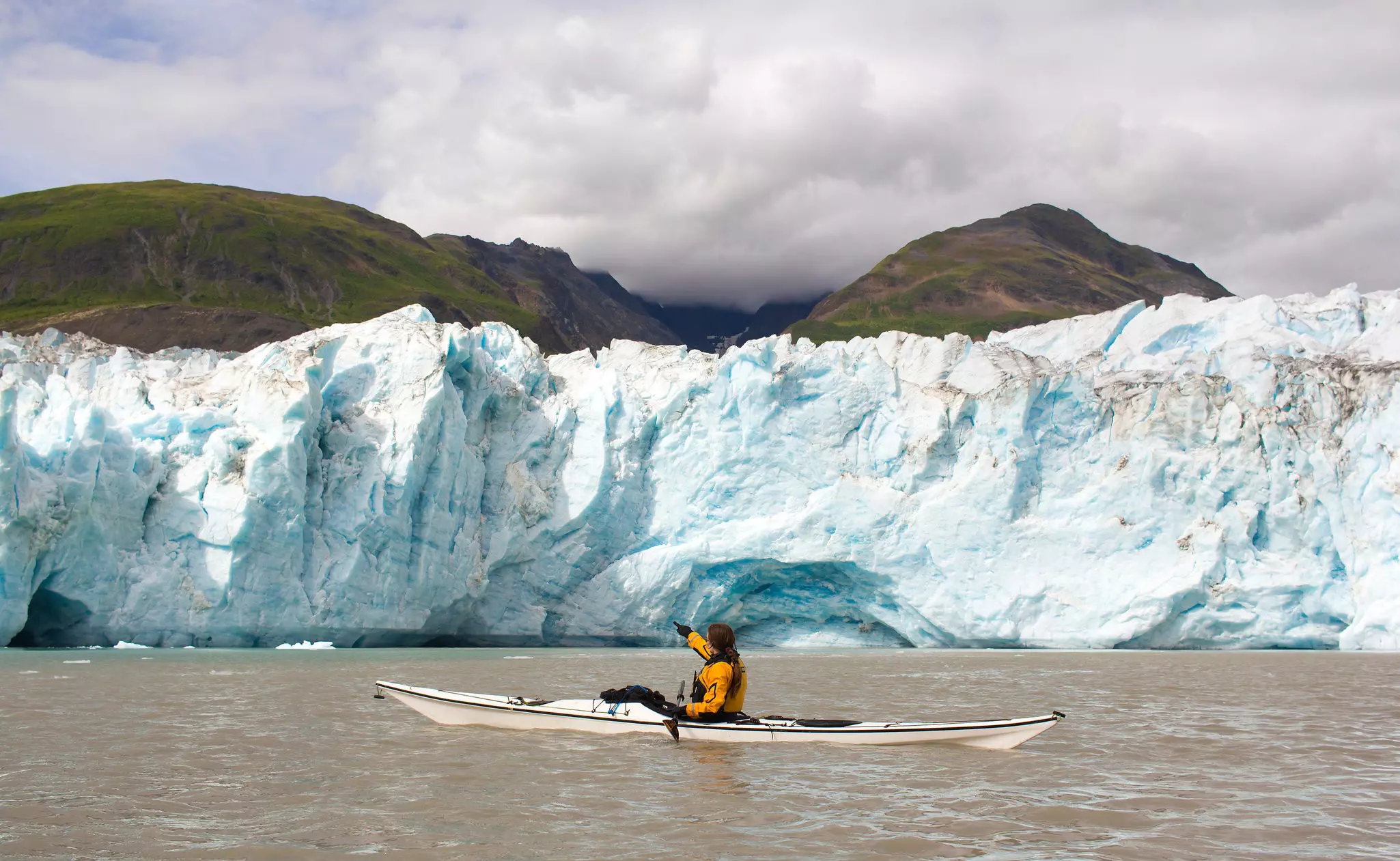 A woman in a yellow kayak paddles through a bay adjacent to a vast wall of ice where a glacier meets the ocean.