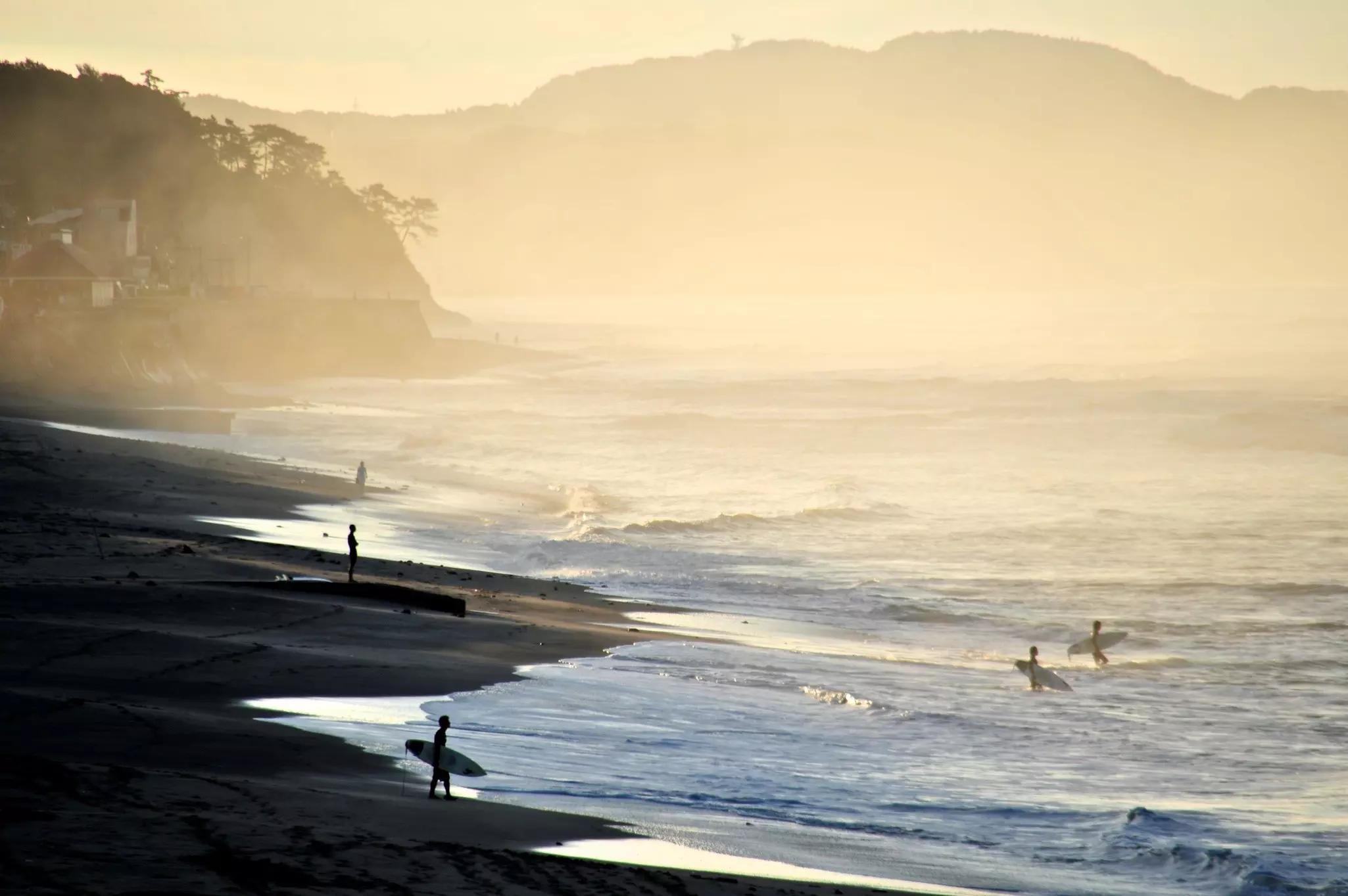 Surfers at a beach are silhouetted in the hazy light of sunset.
