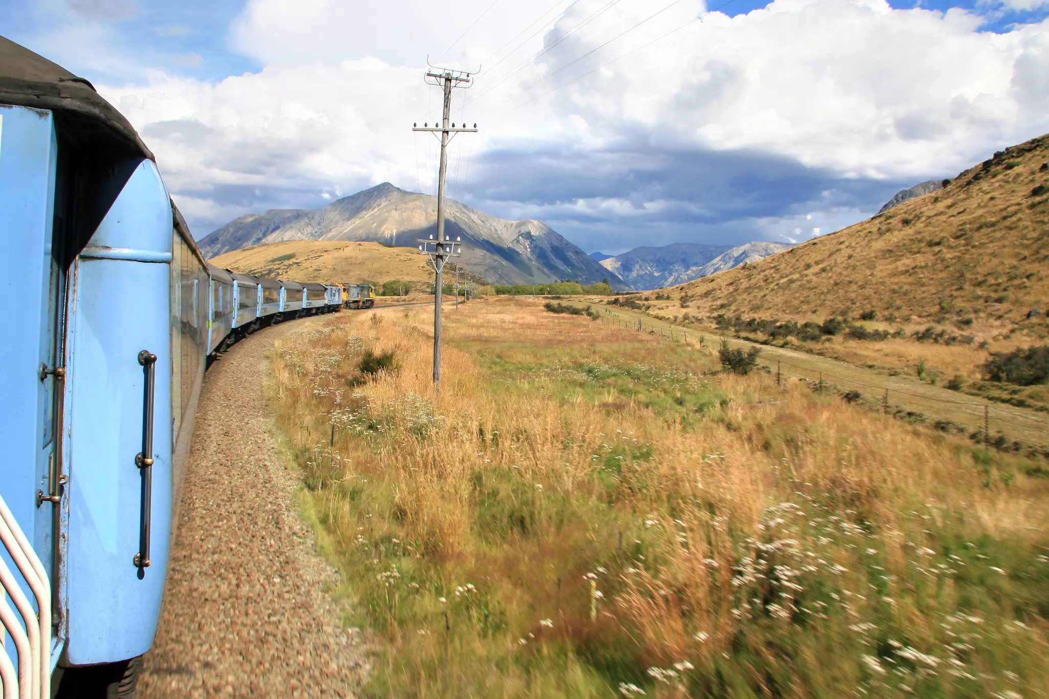 A train with blue carriages follows a curve in the tracks in a mountainous region of New Zealand.