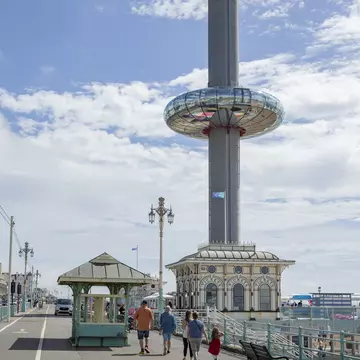 A circular glass observation pod is ascending up a high tower on a beach promenade