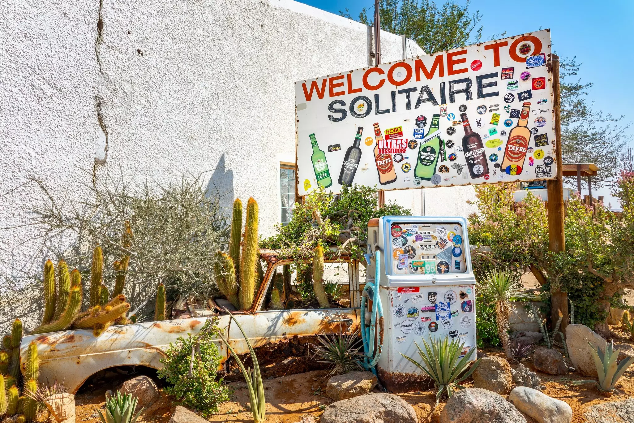A sign welcoming drivers to a town rises above an abandoned gas-station pub and rusting car chassis on the side of a road.