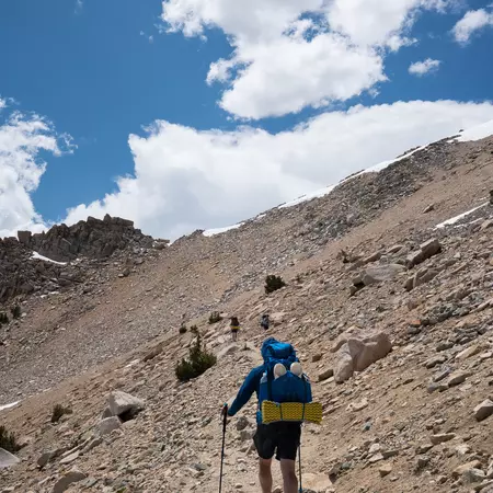 A hiker, carrying a backpack and using hiking poles, follows a rocky mountain trail.