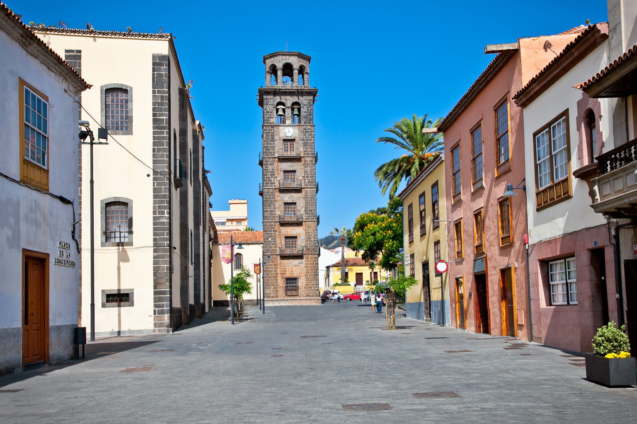 Tall stone bell tower at the end of a pedestrian street lined with pastel colonial houses and a palm tree