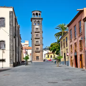 Tall stone bell tower at the end of a pedestrian street lined with pastel colonial houses and a palm tree