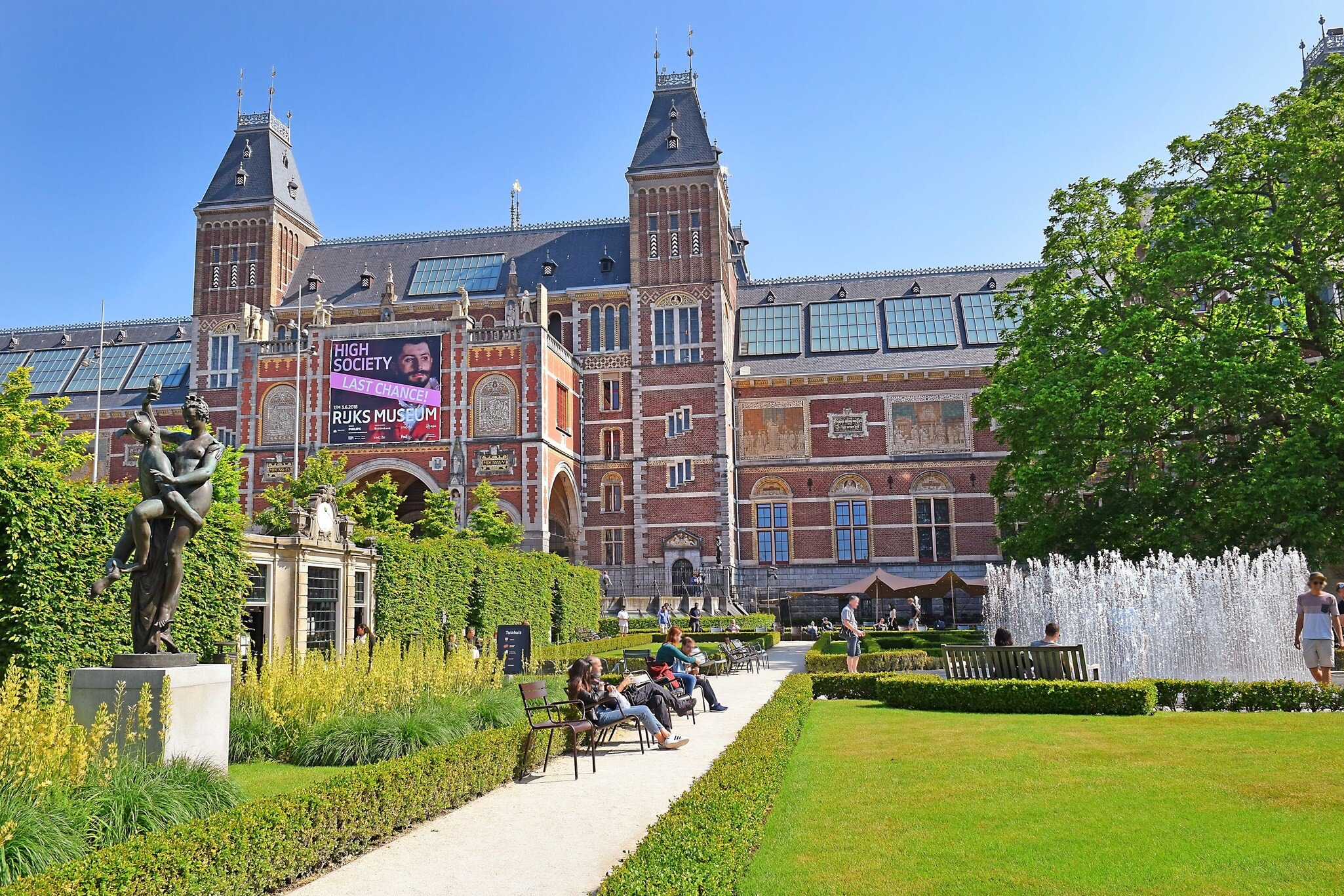 Exterior view of the Rijksmuseum on a sunny day in Amsterdam, with visitors around the fountain at the garden in the foreground
