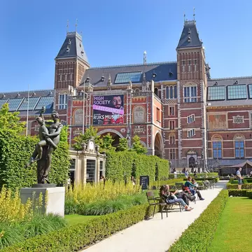 Exterior view of the Rijksmuseum on a sunny day in Amsterdam, with visitors around the fountain at the garden in the foreground
