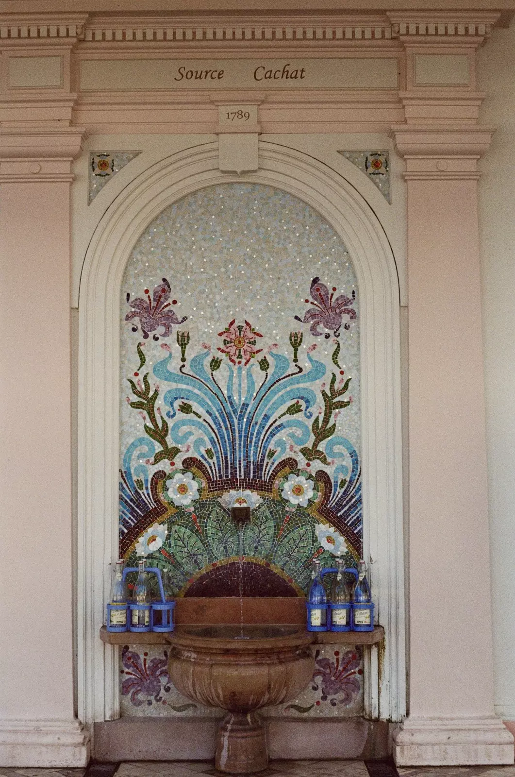 A spring backed by a colorful mosaic with water bottles on either side waiting to be filled.
