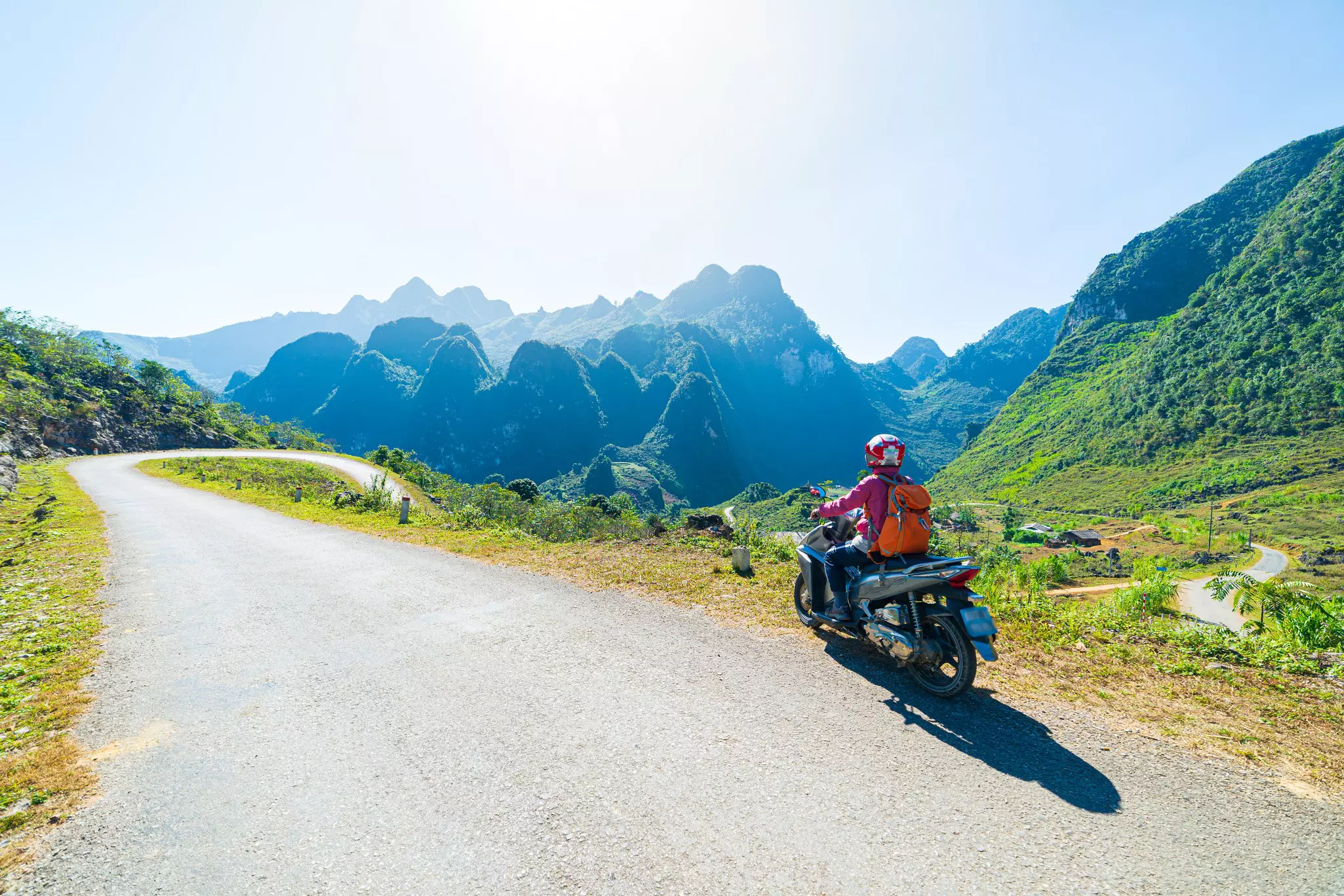 A man on a motor scooter rides on windy mountain road. Hazy green hills are in the distance.