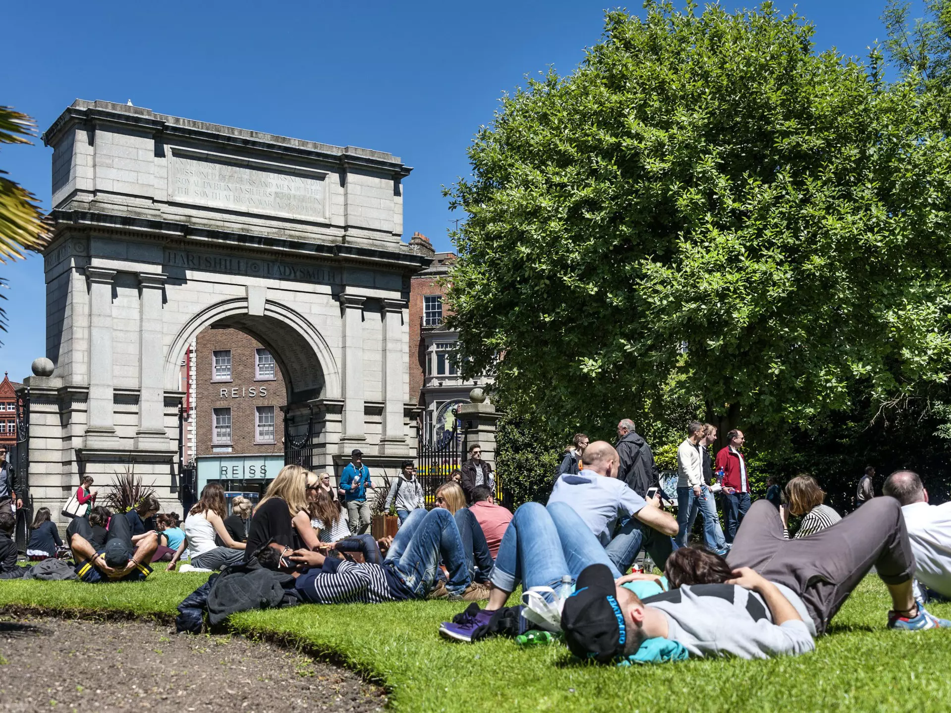People lie on the grass in a city park next to a stone arch.