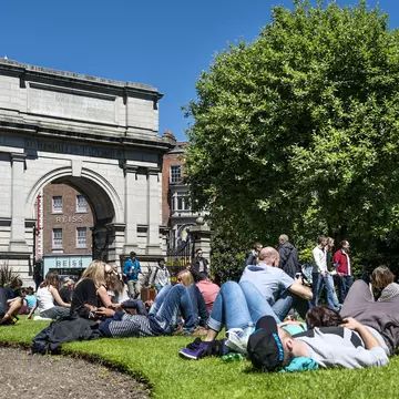 People lie on the grass in a city park next to a stone arch.