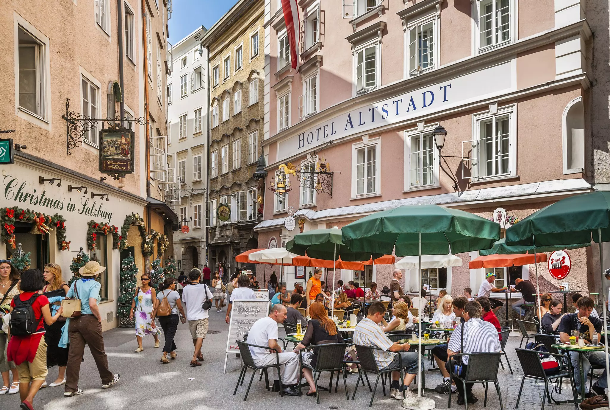 Diners at outdoor tables under umbrellas, with others walking on the street in a plaza enclosed by historic commercial buildings on a summer day.