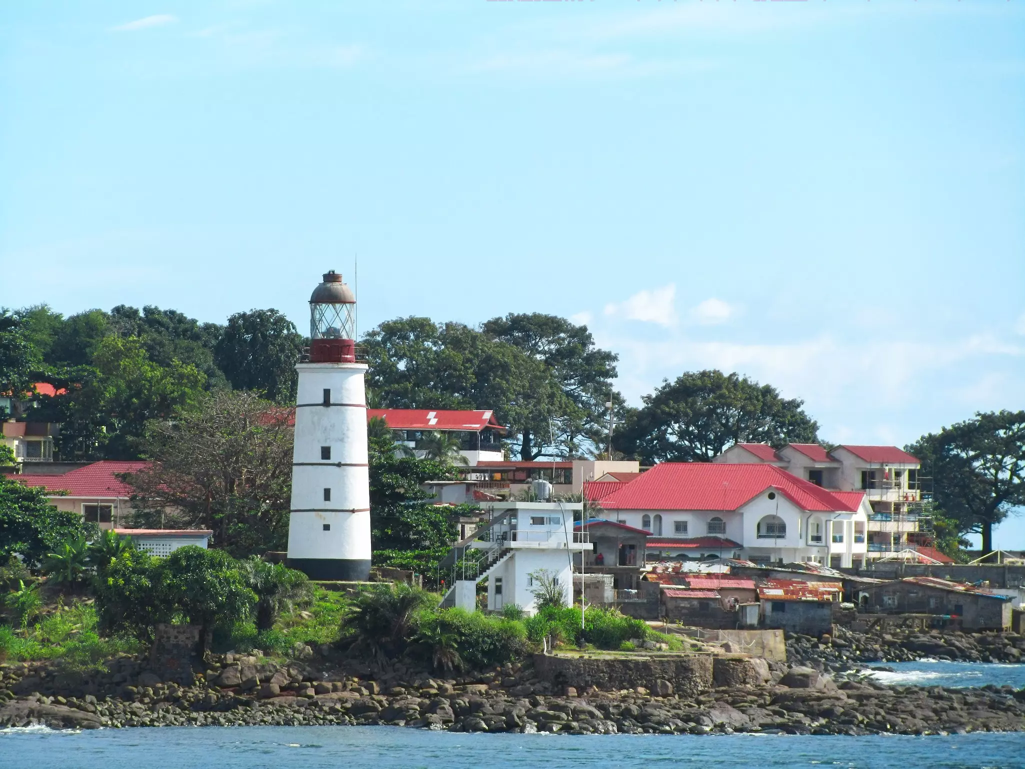 A view from the water of a white-painted lighthouse and other buildings on the shore.
