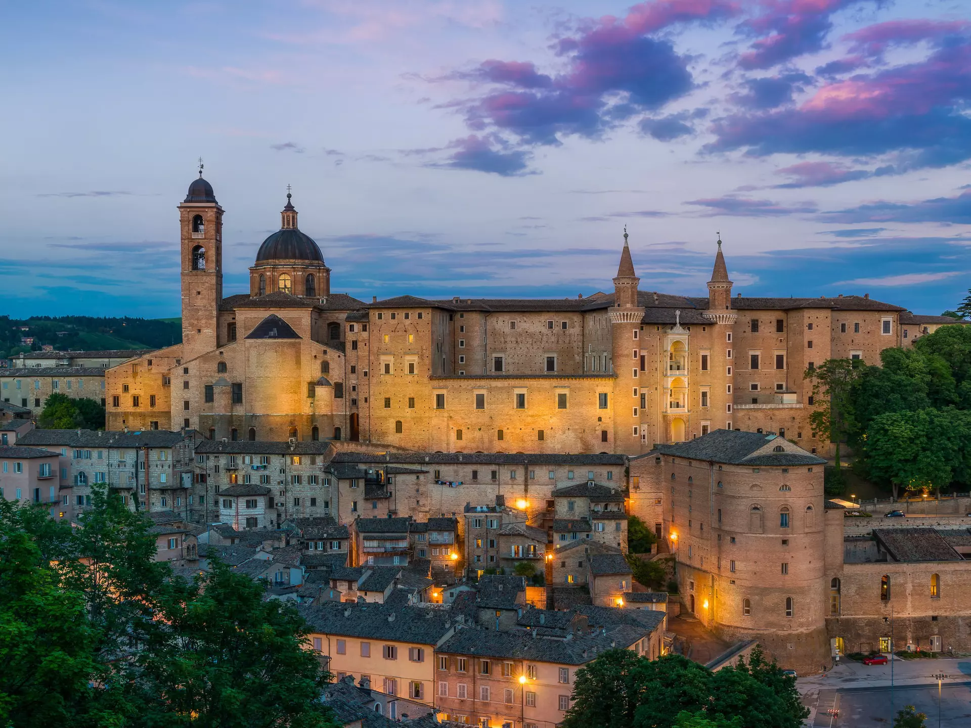 A view of Ducal Palace in Urbino at sunset; the sky is blue and purple and the palace is lit from below. Urbino, Le Marche, Italy.