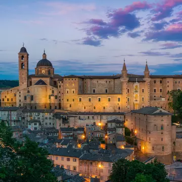A view of Ducal Palace in Urbino at sunset; the sky is blue and purple and the palace is lit from below. Urbino, Le Marche, Italy.