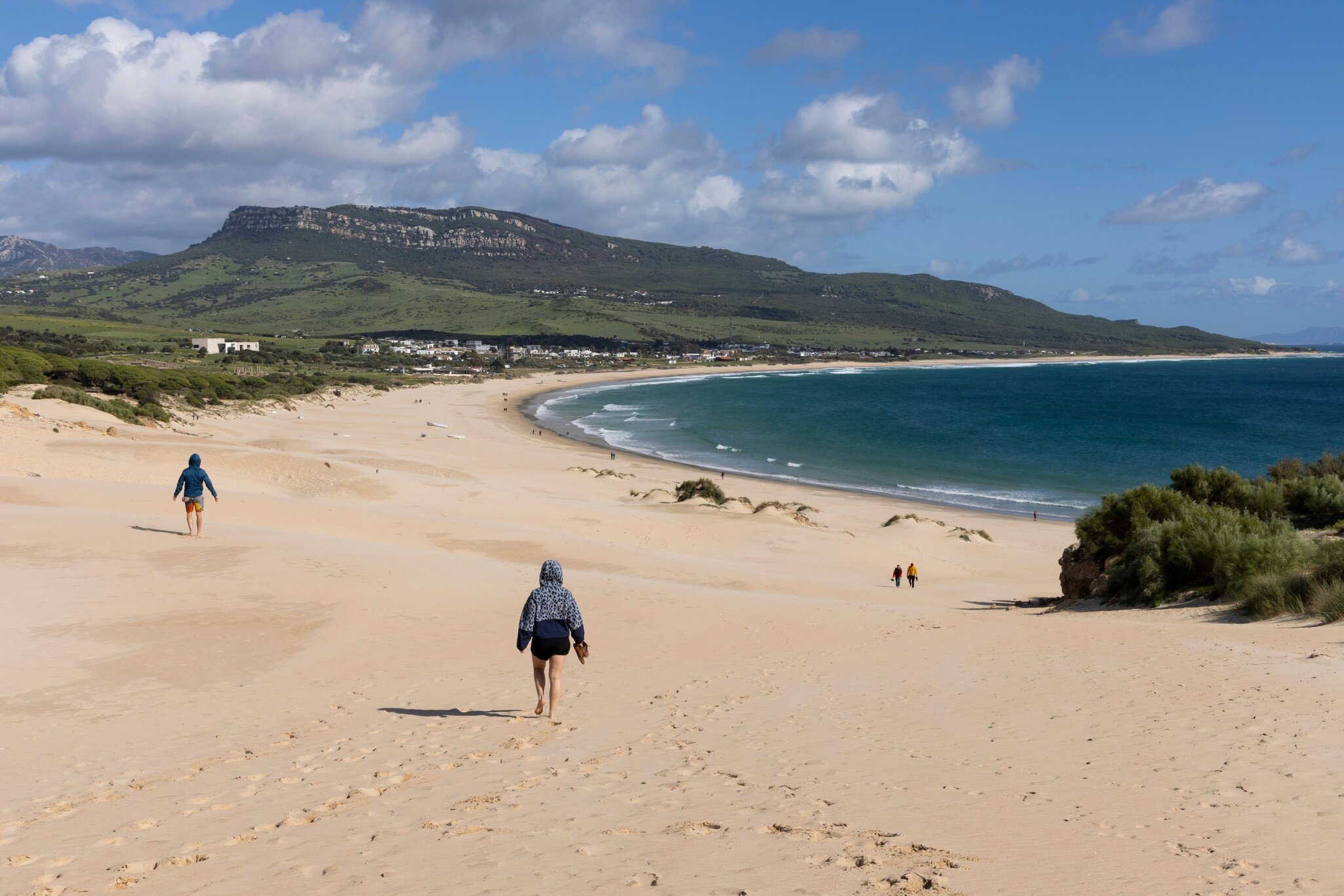 The backs of two people walking separately with two others in the distance on a wide sandy beach. The ocean is to the right and green-covered mountains are in the distance.