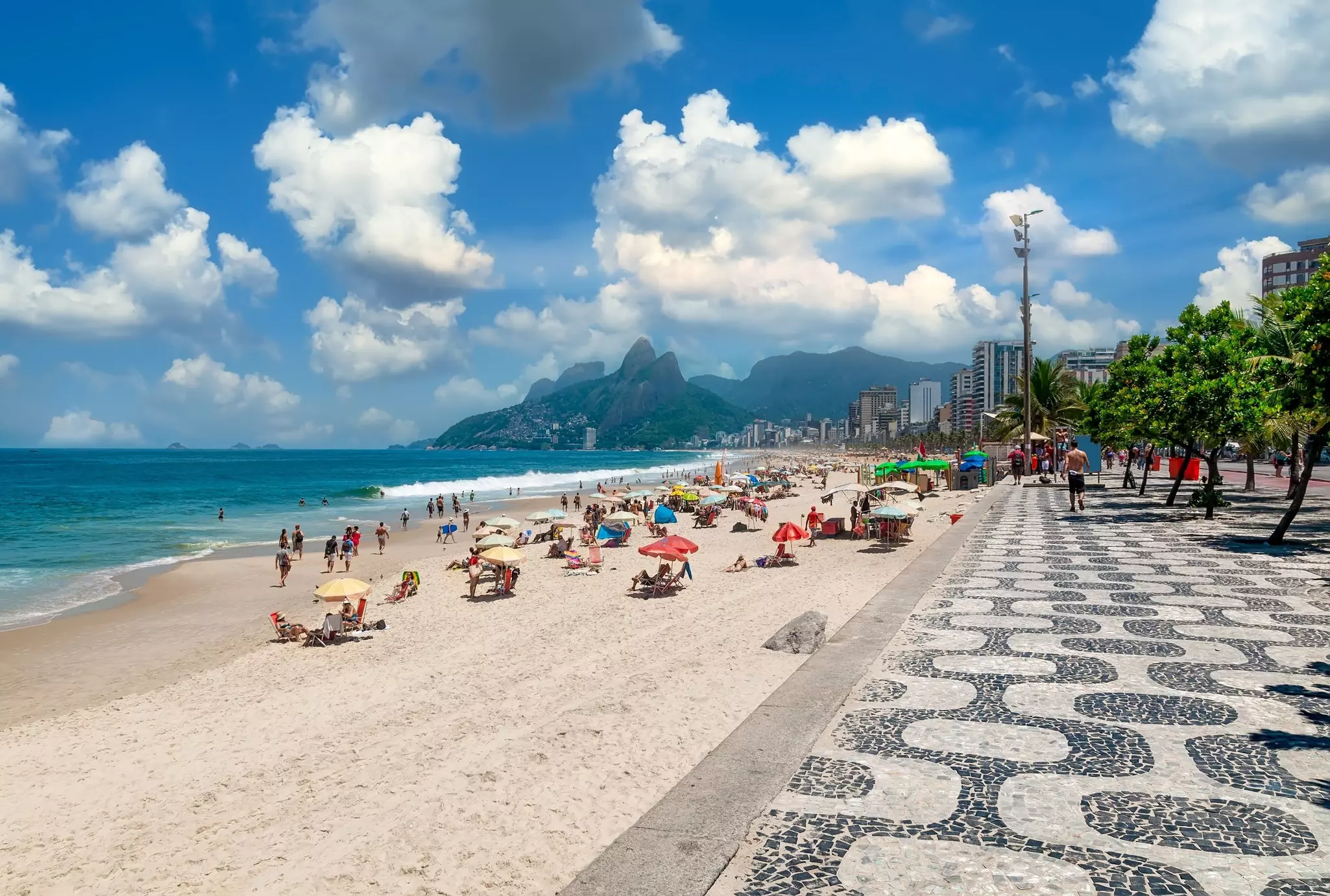 A black-and-white mosaic sidewalk runs beside a sandy beach where people sit under multicolored umbrellas; peaked mountains are at the far shore.