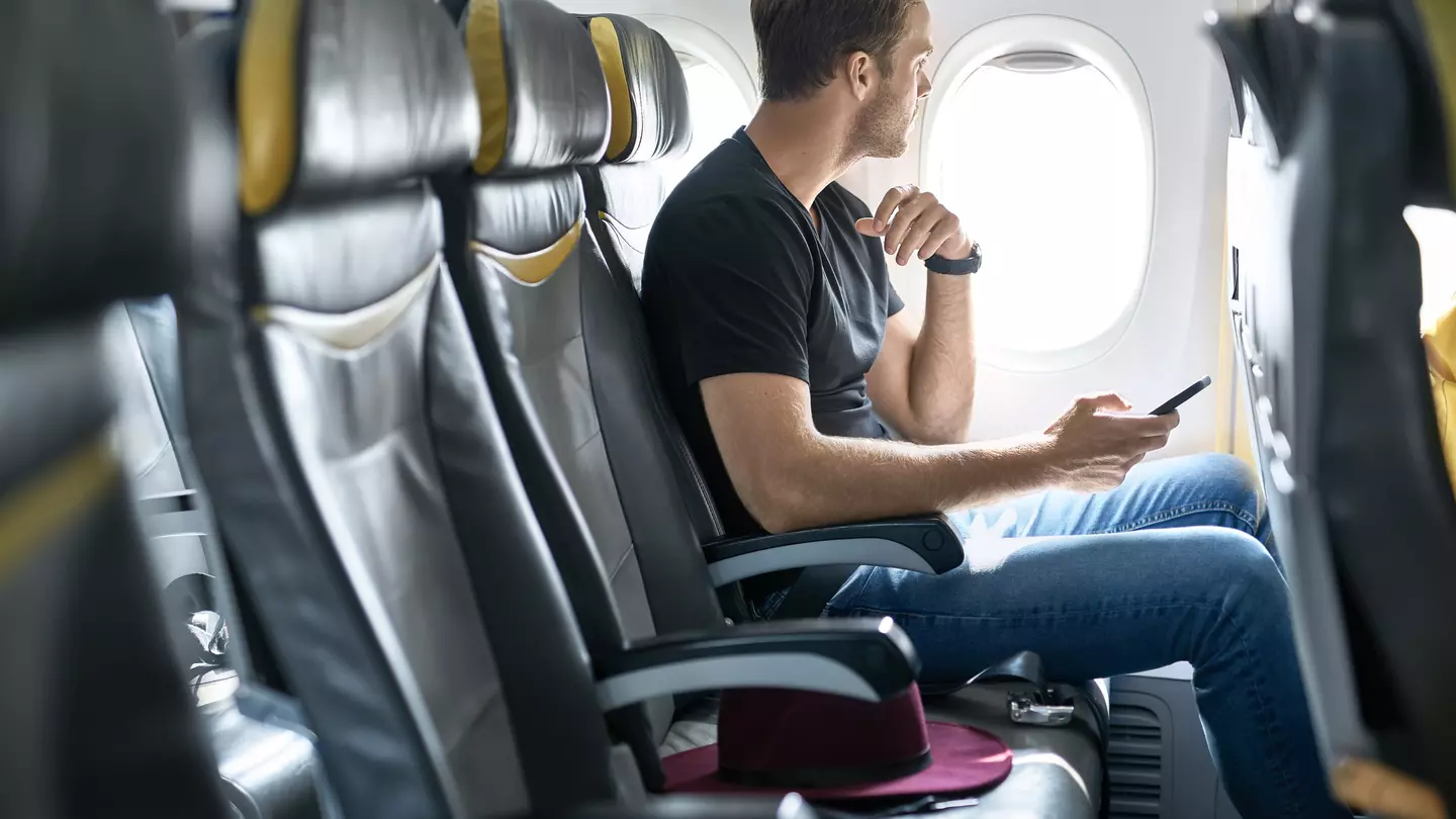 A young male passenger sits in an airplane seat next to the window with a mobile phone