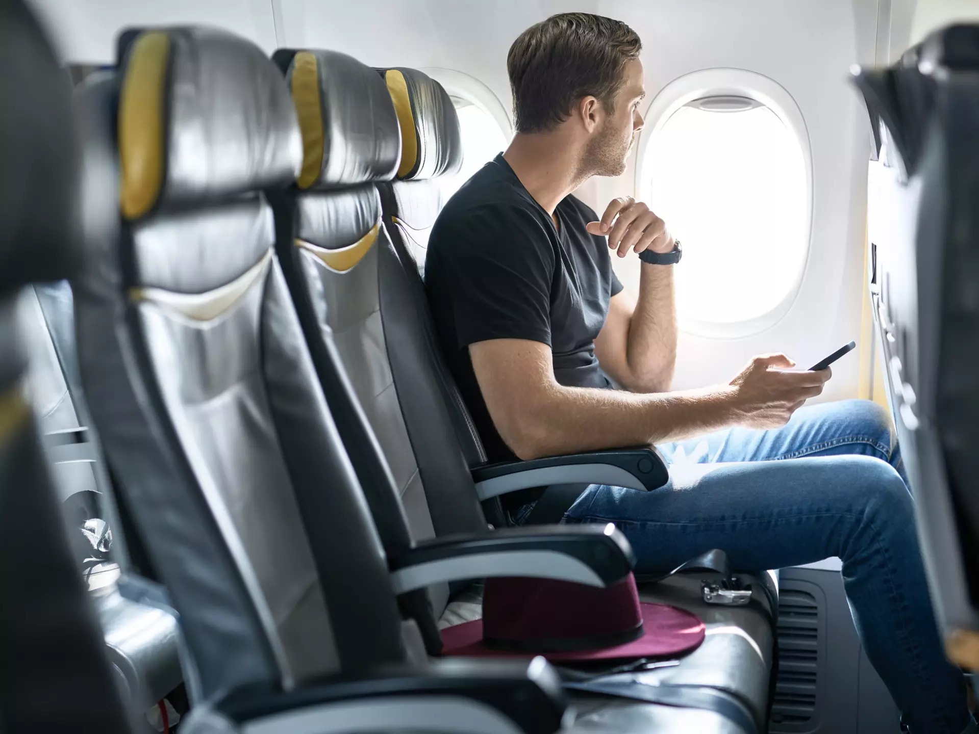 A young male passenger sits in an airplane seat next to the window with a mobile phone