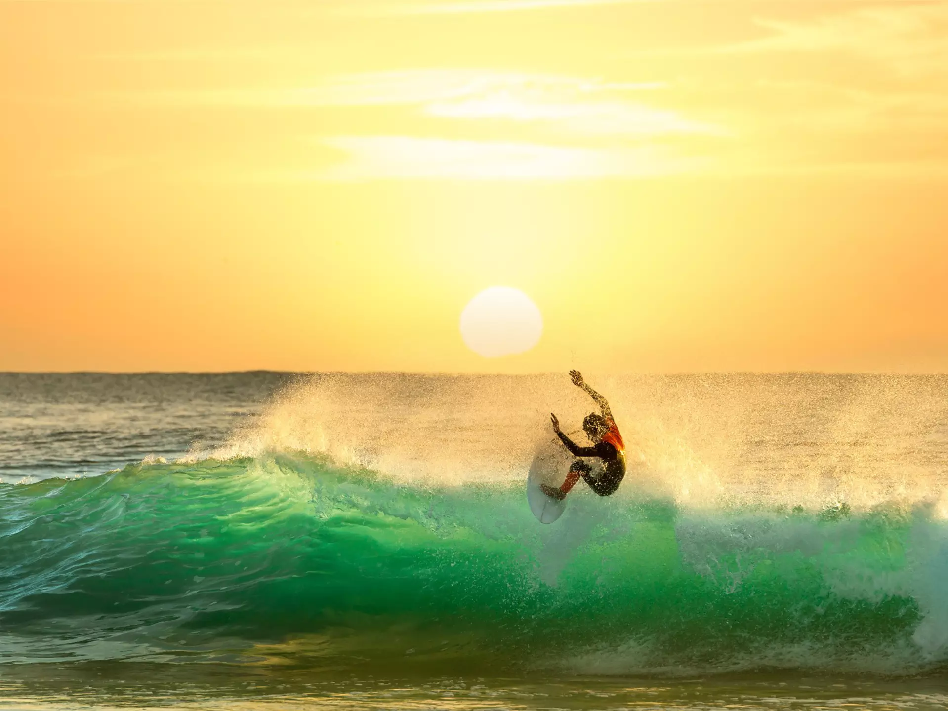 A surfer atop the crest of a wave almost floats in the air; the wave, lit by a sun right on the horizon, is glowing green. The sky is a brilliant gold colour.
