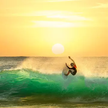 A surfer atop the crest of a wave almost floats in the air; the wave, lit by a sun right on the horizon, is glowing green. The sky is a brilliant gold colour.