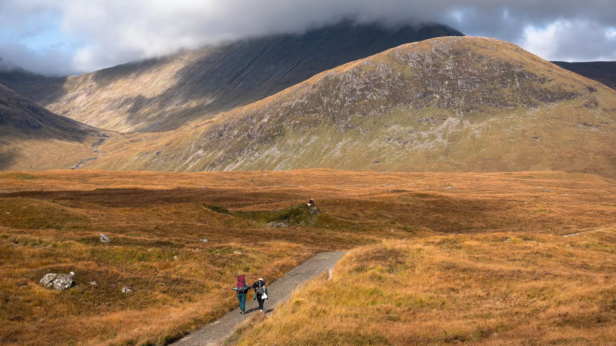 Two hikers are seen from behind on a trail through meadows, with cloud-covers hills in the distance.