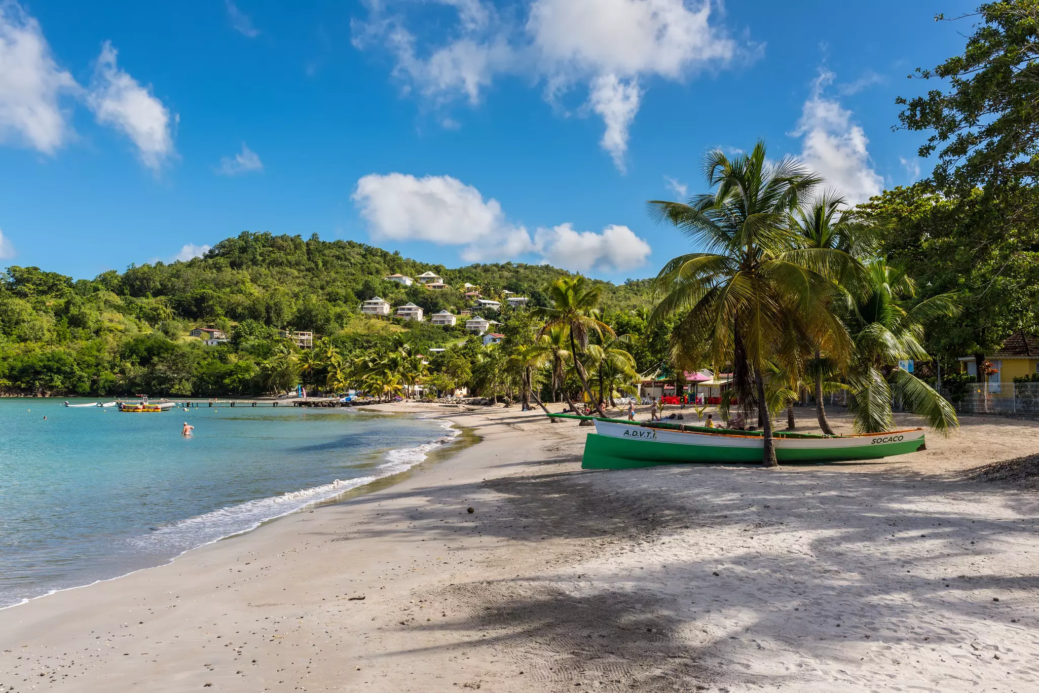 A green boat sits on a a beach surrounded by palm trees with villas in the hills beyond