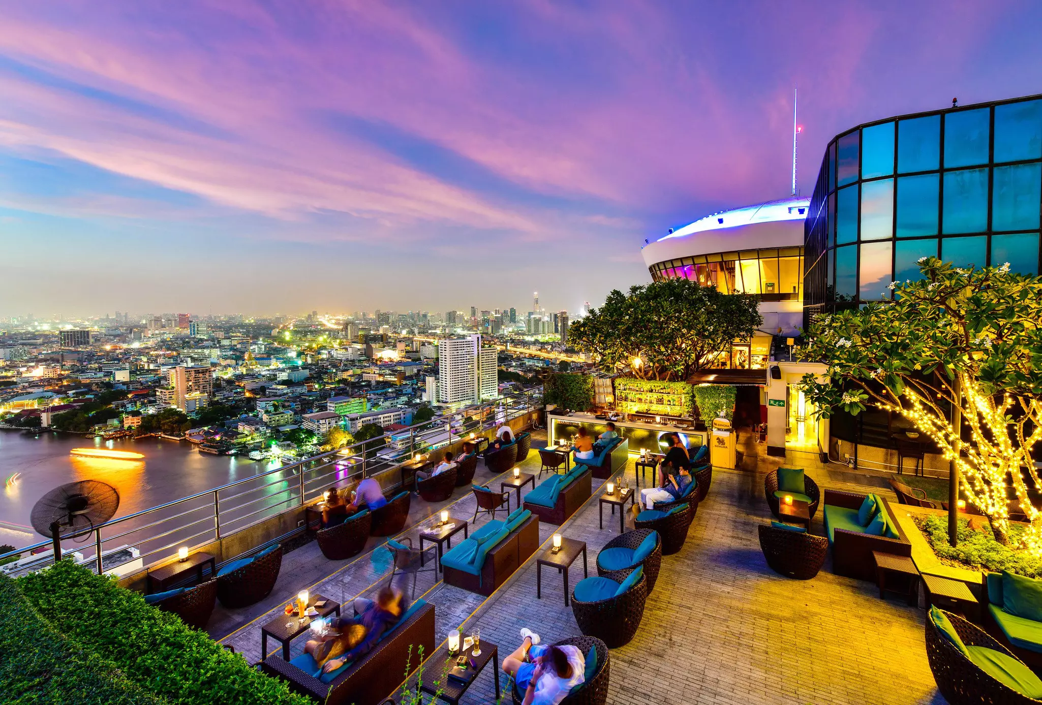 Diners and drinkers relaxing on lounge chairs on a rooftop restaurant with views over the river and city below.