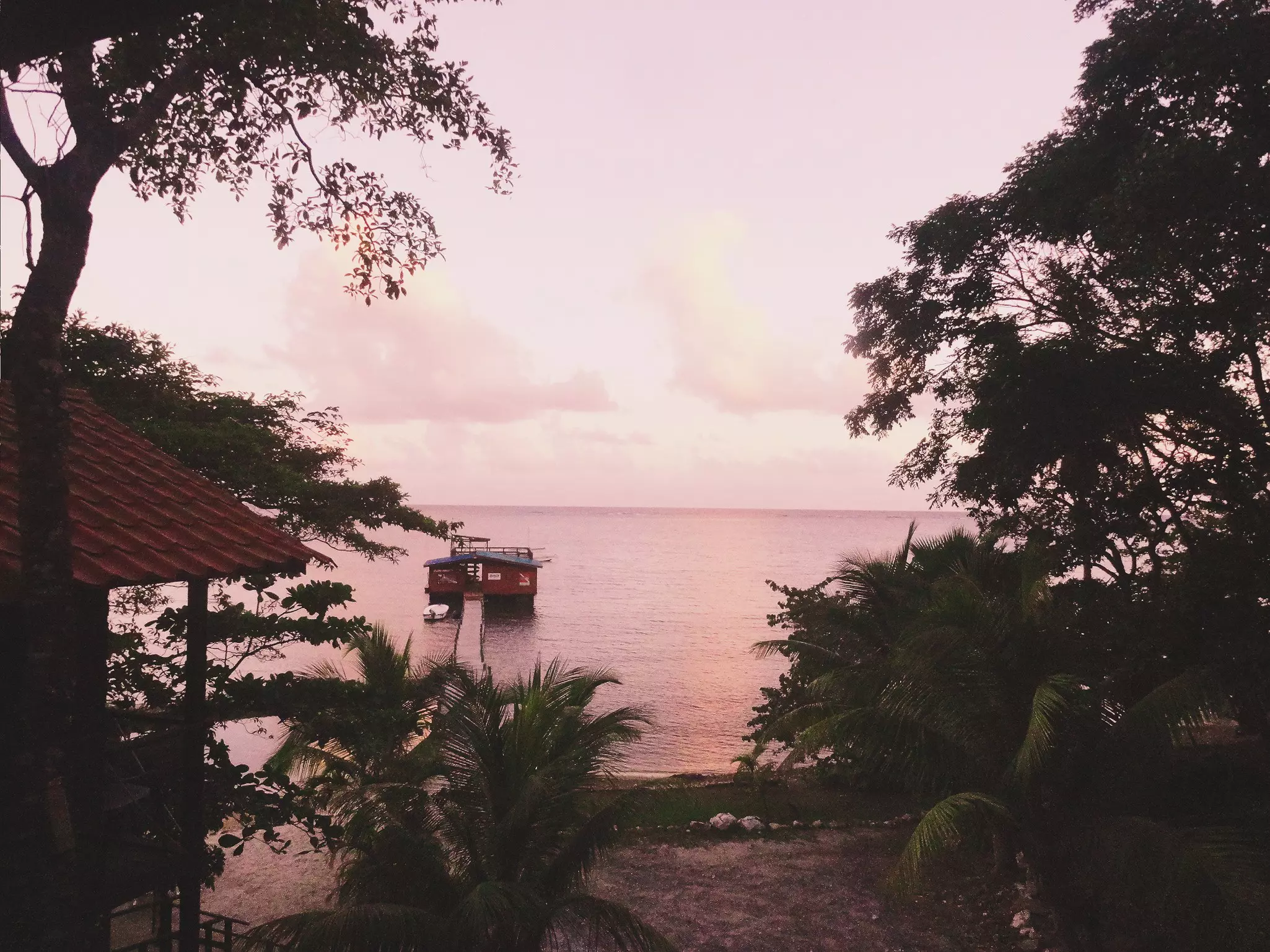 A view out toward a beach, a pier, and the water beyond at sunset. The scene appears pink from the light.