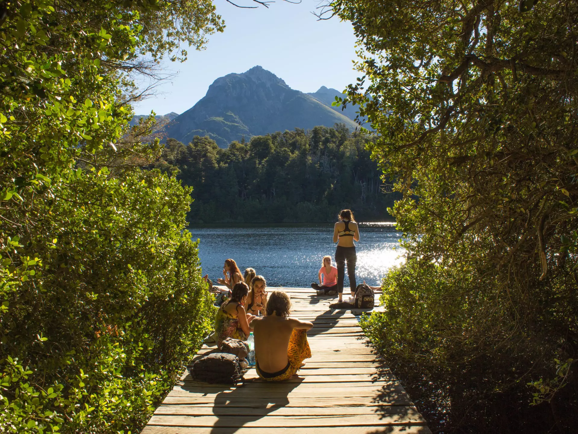 People on a dock by a lake in sunshine; greenery lines either side of the dock.