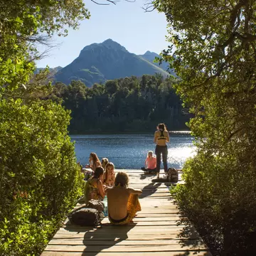 People on a dock by a lake in sunshine; greenery lines either side of the dock.